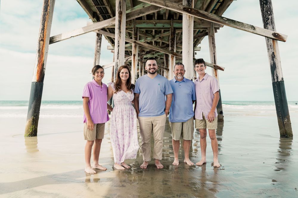 Family of five smiling together under a wooden beach pier on a cloudy day at the shore.