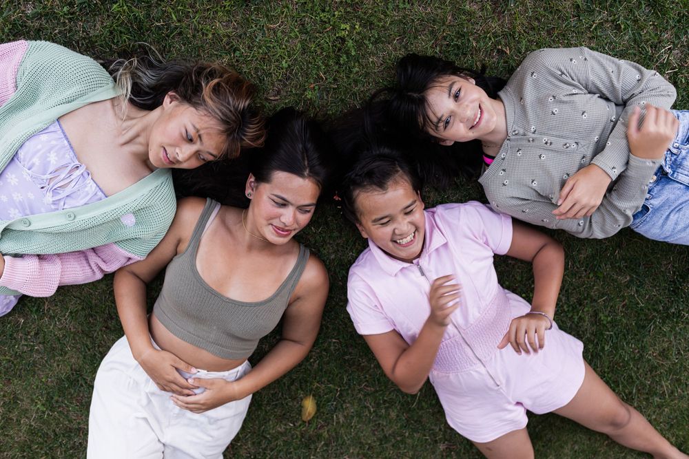 Five young women lying on green grass in a circle, smiling and laughing together outdoors.