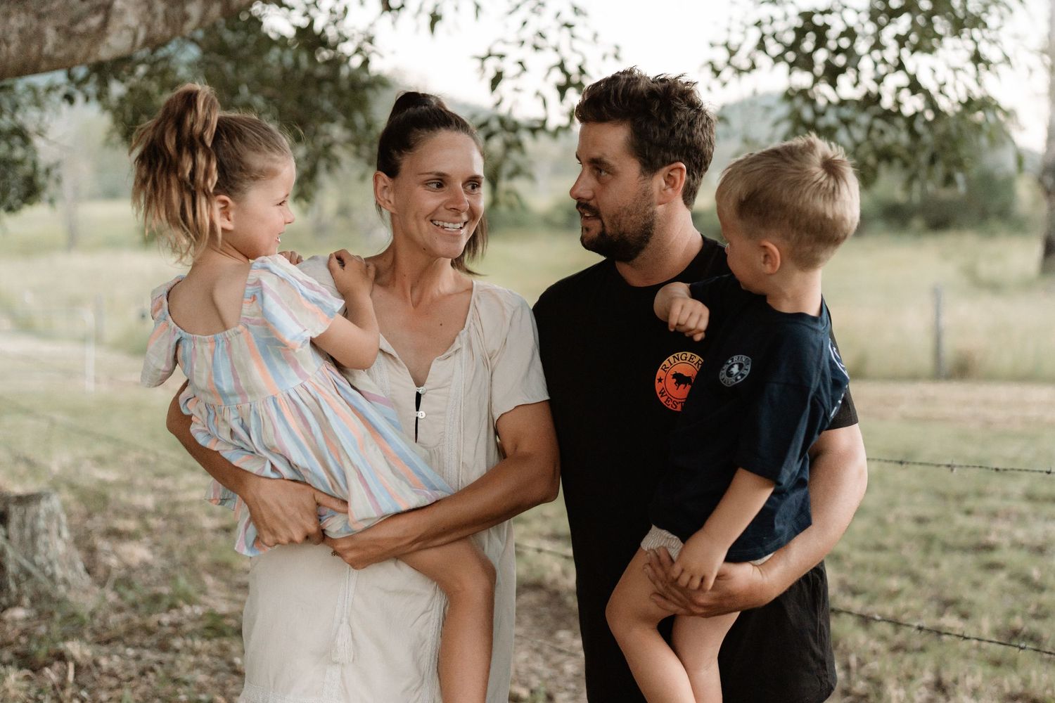 A happy family of four poses outdoors, with parents holding two young children in a lush green countryside setting.