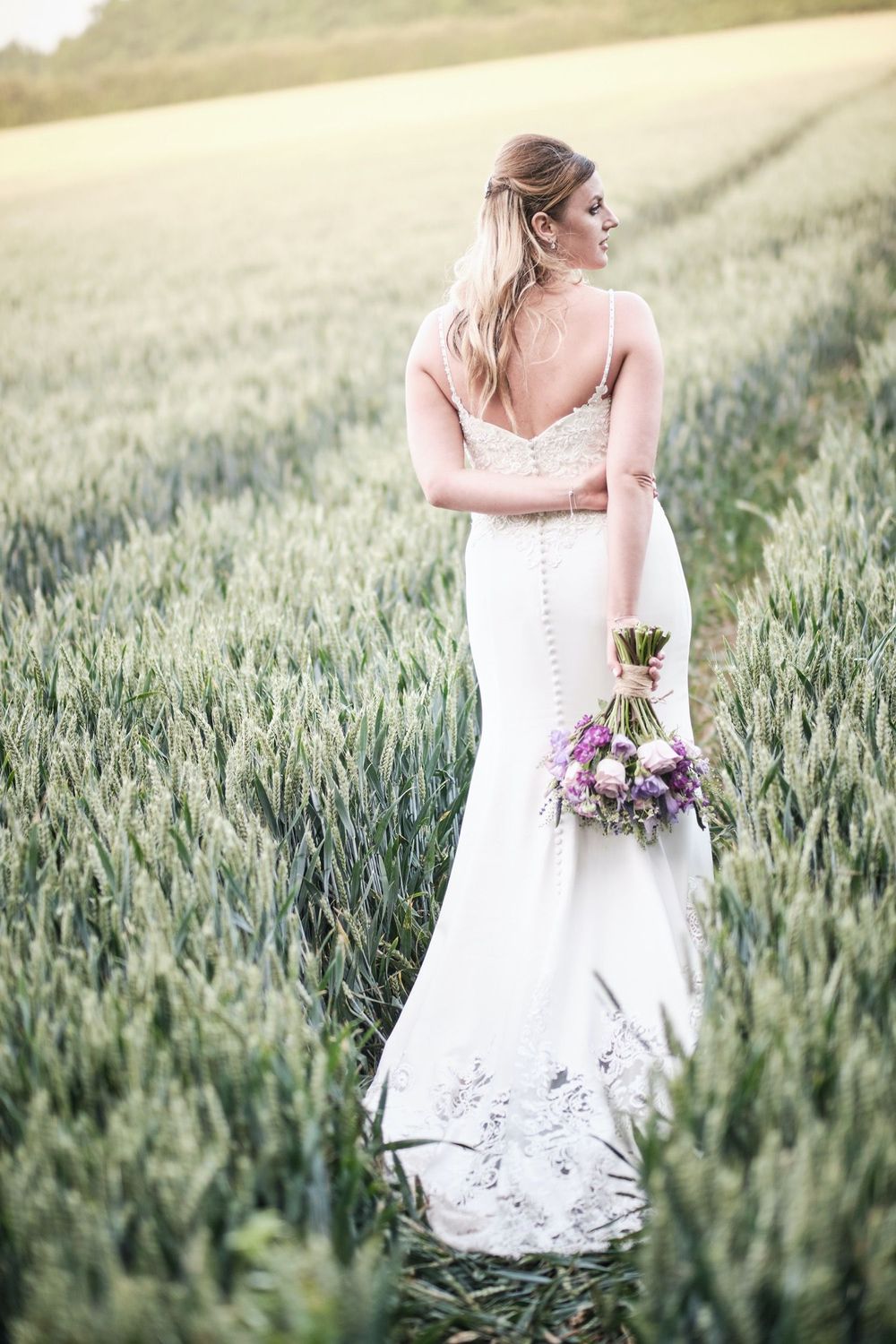 A bride in a white lace wedding dress stands in a green wheat field holding a purple bouquet at sunset.