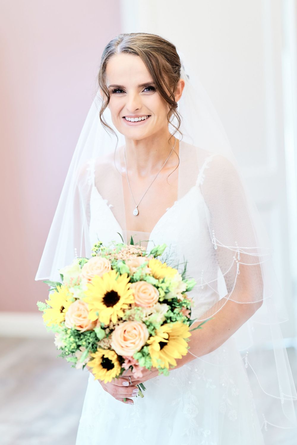 A bride in white dress holds a vibrant sunflower and rose bouquet while smiling.