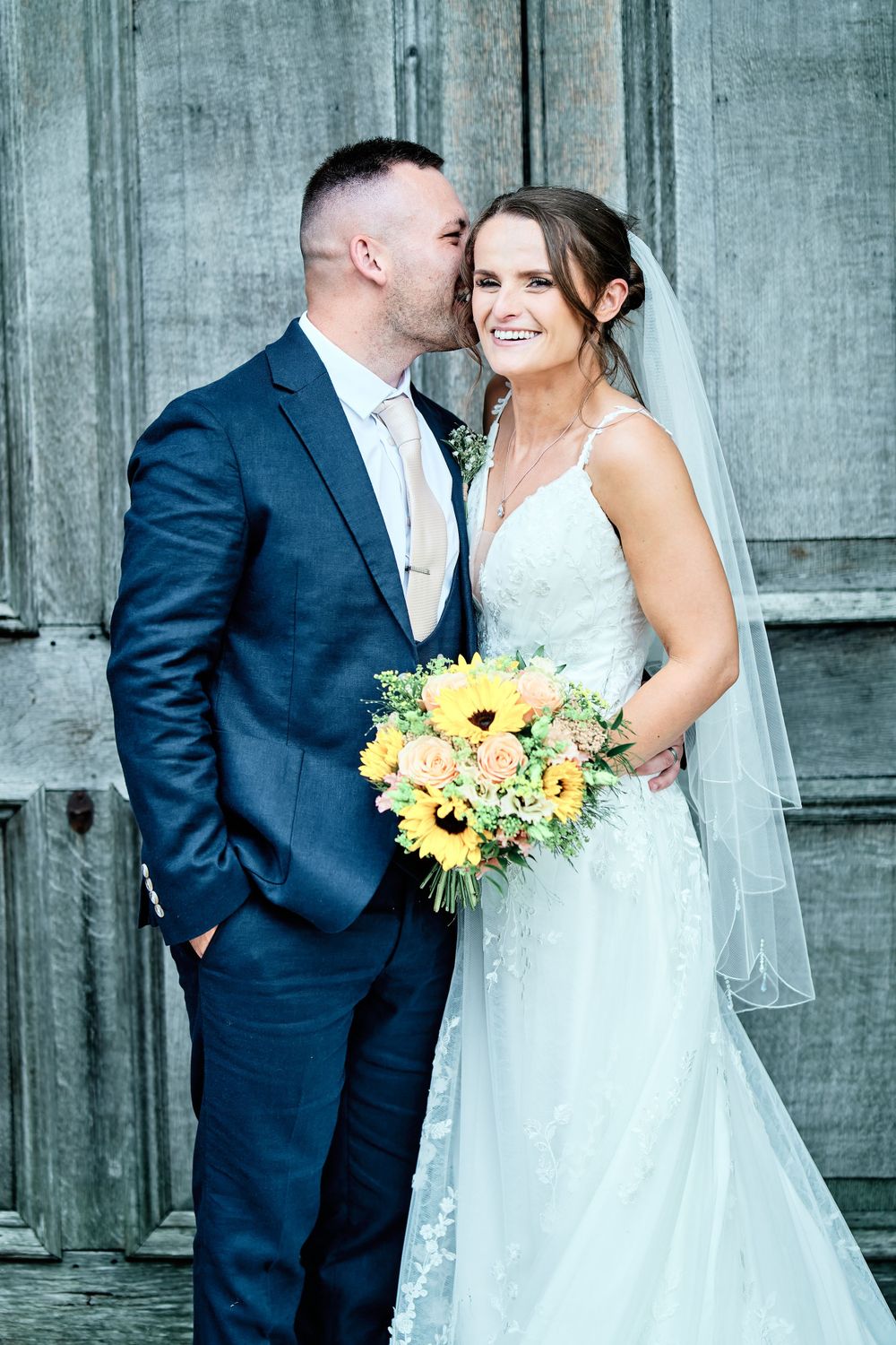 Intimate wedding moment captured against rustic wooden backdrop with sunflower bouquet.