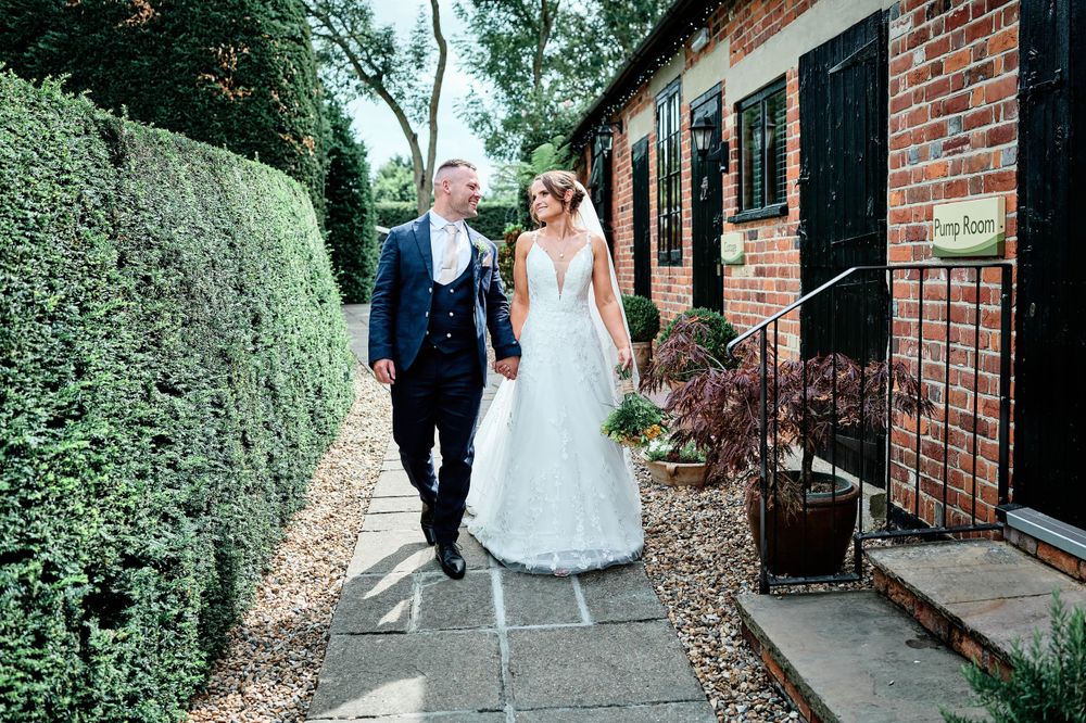 Wedding couple walks along stone pathway beside brick building with manicured hedges.