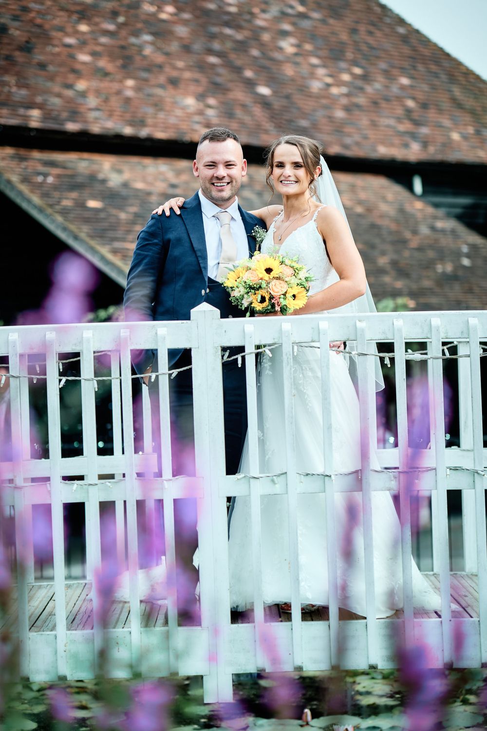 A wedding couple stands on a white balcony with purple flowers in the foreground at an outdoor venue.