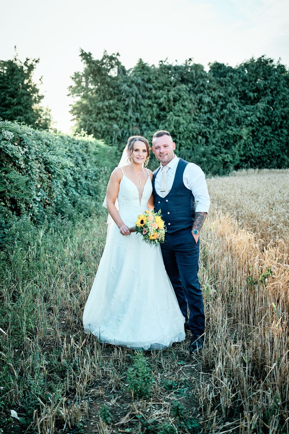 Wedding portrait of couple standing in wheat field against green hedges during sunset.