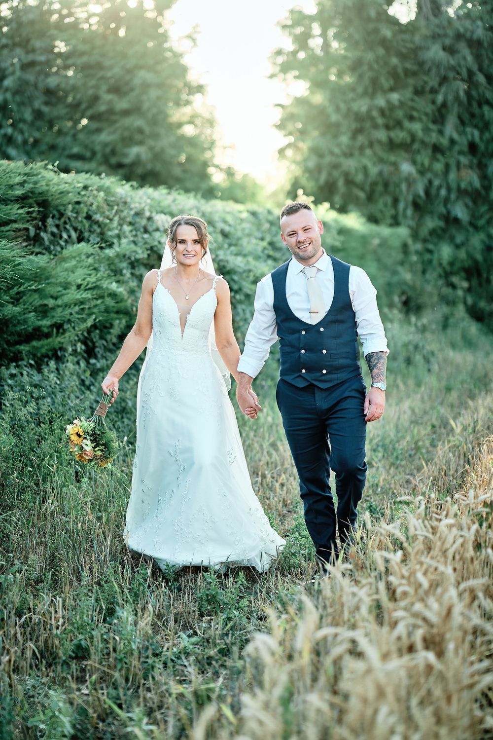 Newlyweds walking through tall grass path with sunlit trees in background.