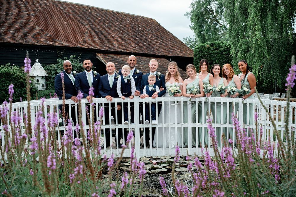 Wedding party poses on a white bridge over a pond with purple flowers blooming in the foreground.