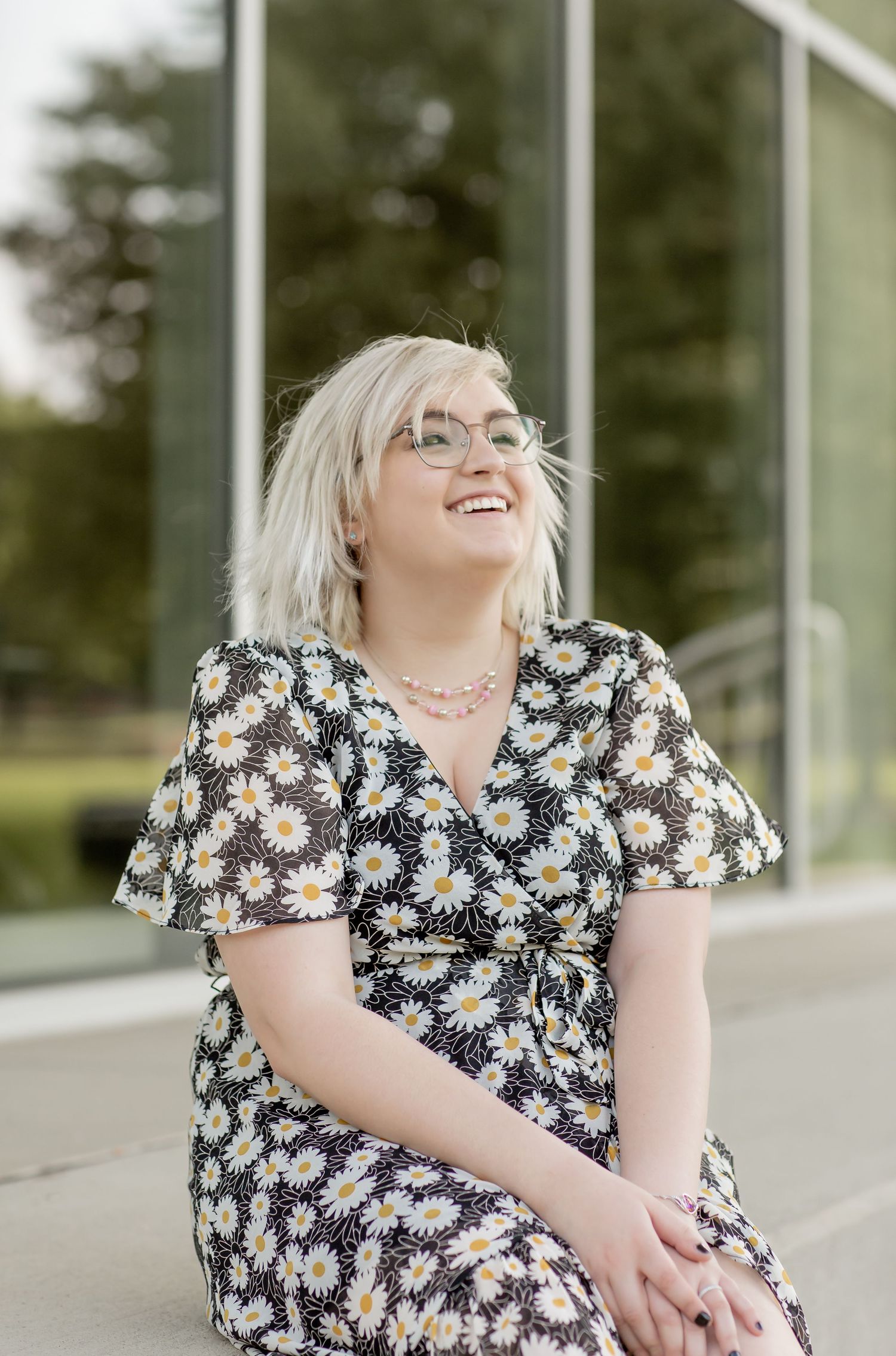 Blonde woman in daisy print dress smiling outdoors wearing glasses.