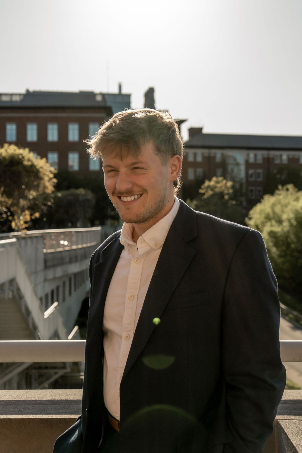 Smiling young man in black blazer standing outdoors on sunny day with brick building in background.