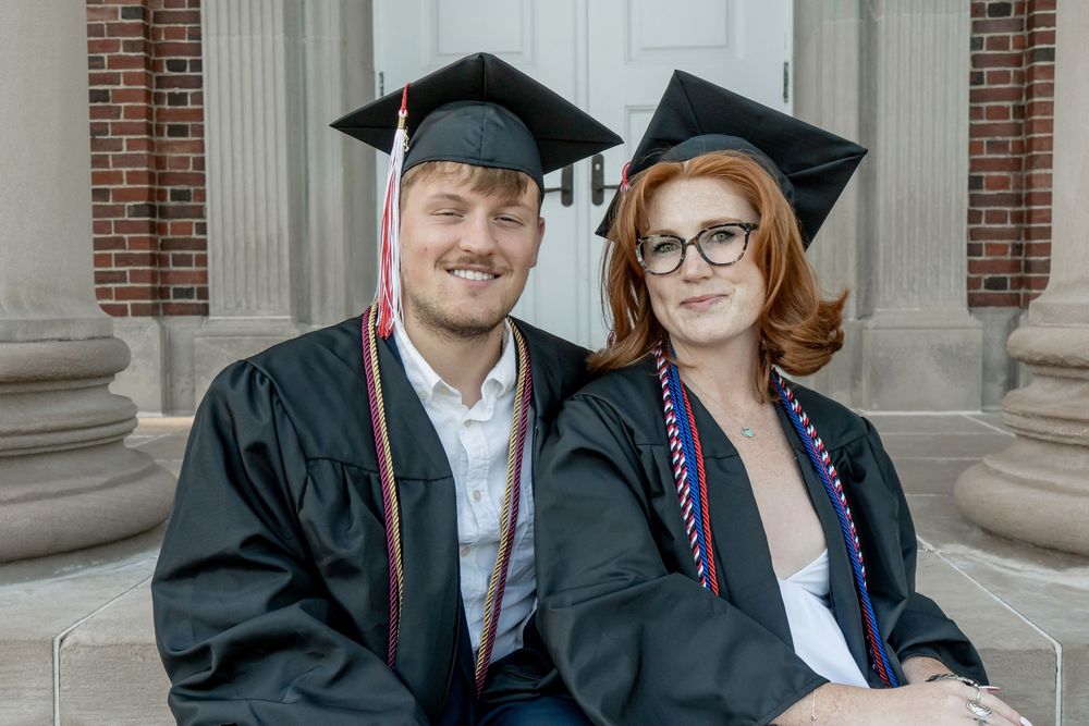 Two smiling graduates in black caps and gowns posing together on steps in front of a brick building.