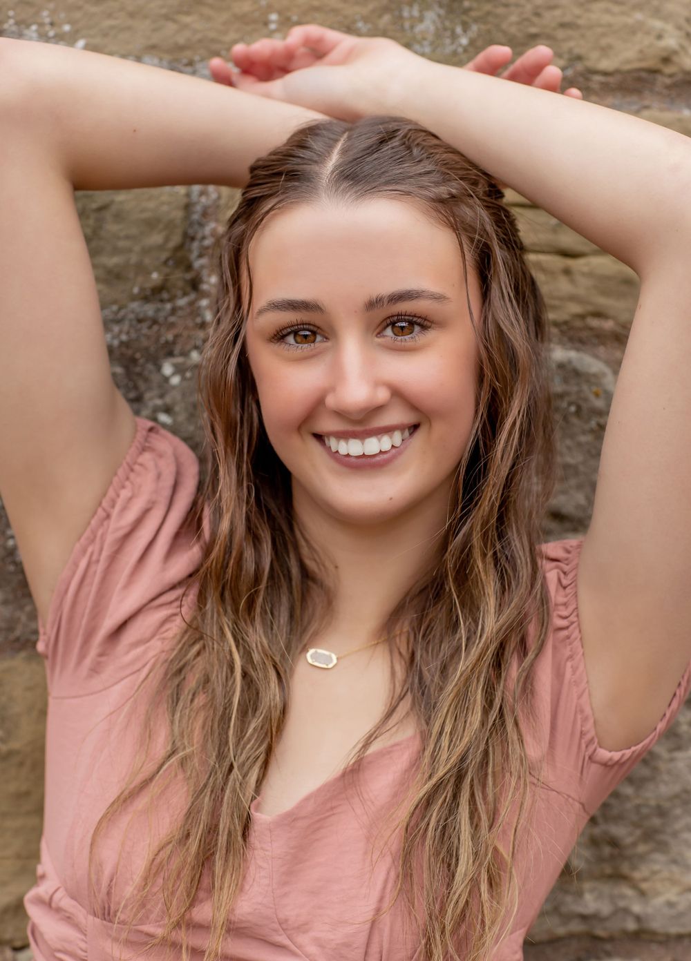 Smiling teenage girl with long highlighted hair wearing a pink top, posing against a stone wall outdoors.