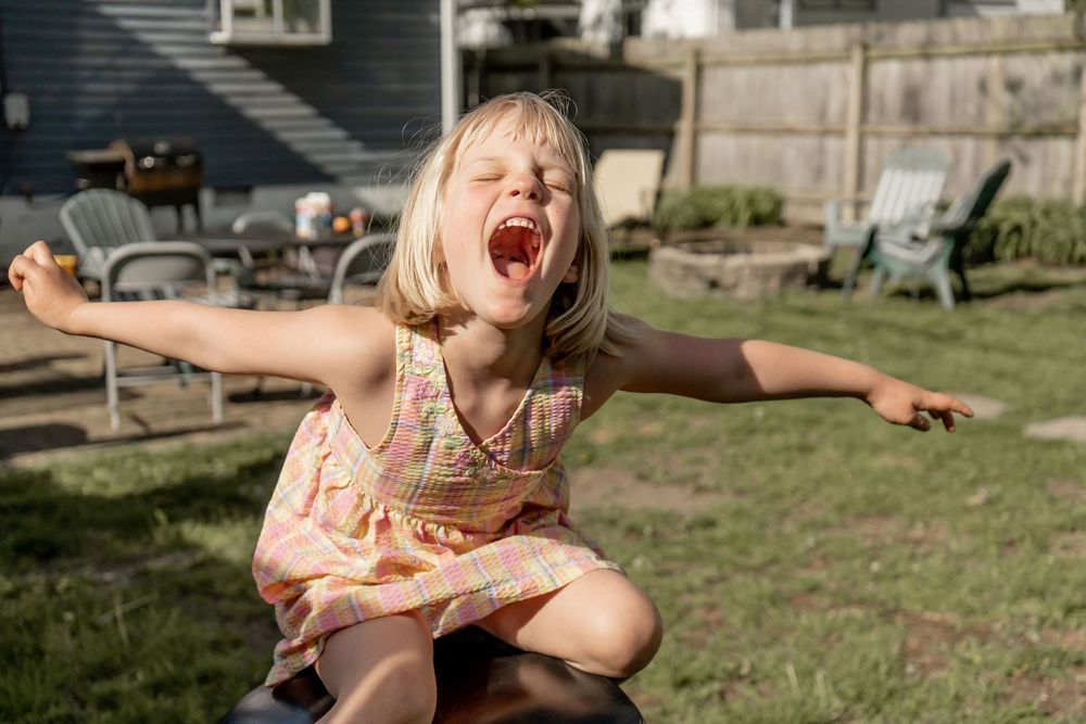 Excited young girl in a floral dress spreading her arms wide and shouting joyfully in a backyard setting.