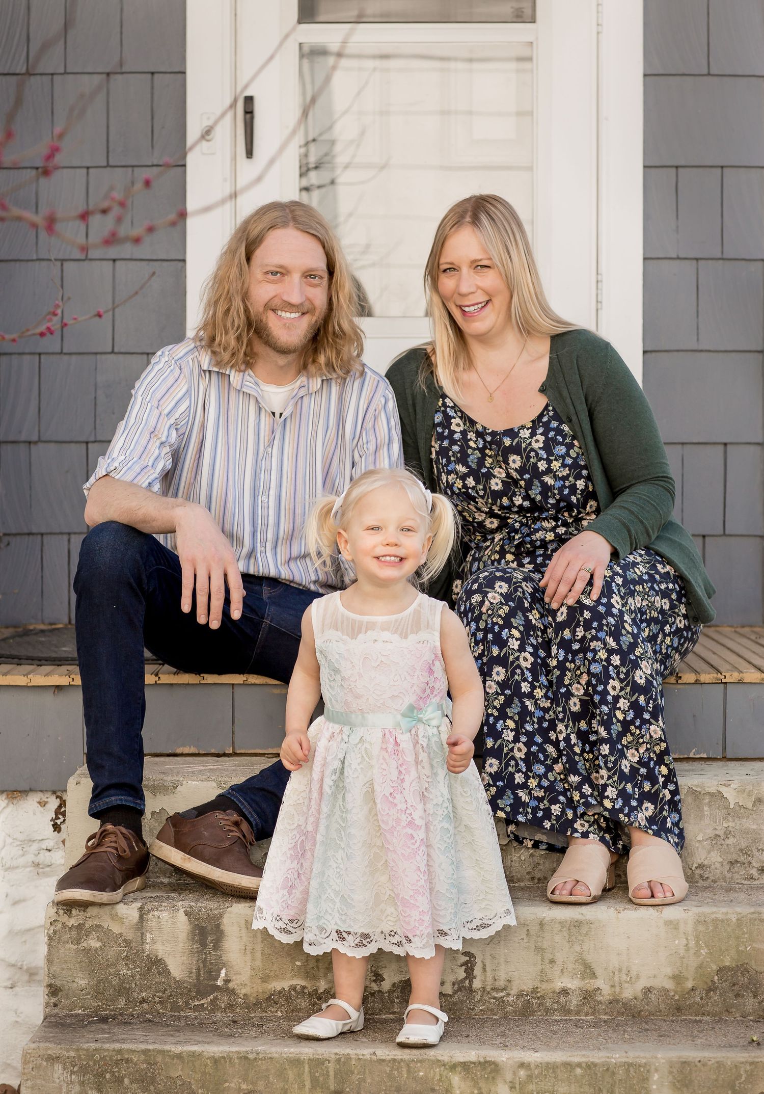 Young family of three posing on home front steps with toddler girl in lace dress.