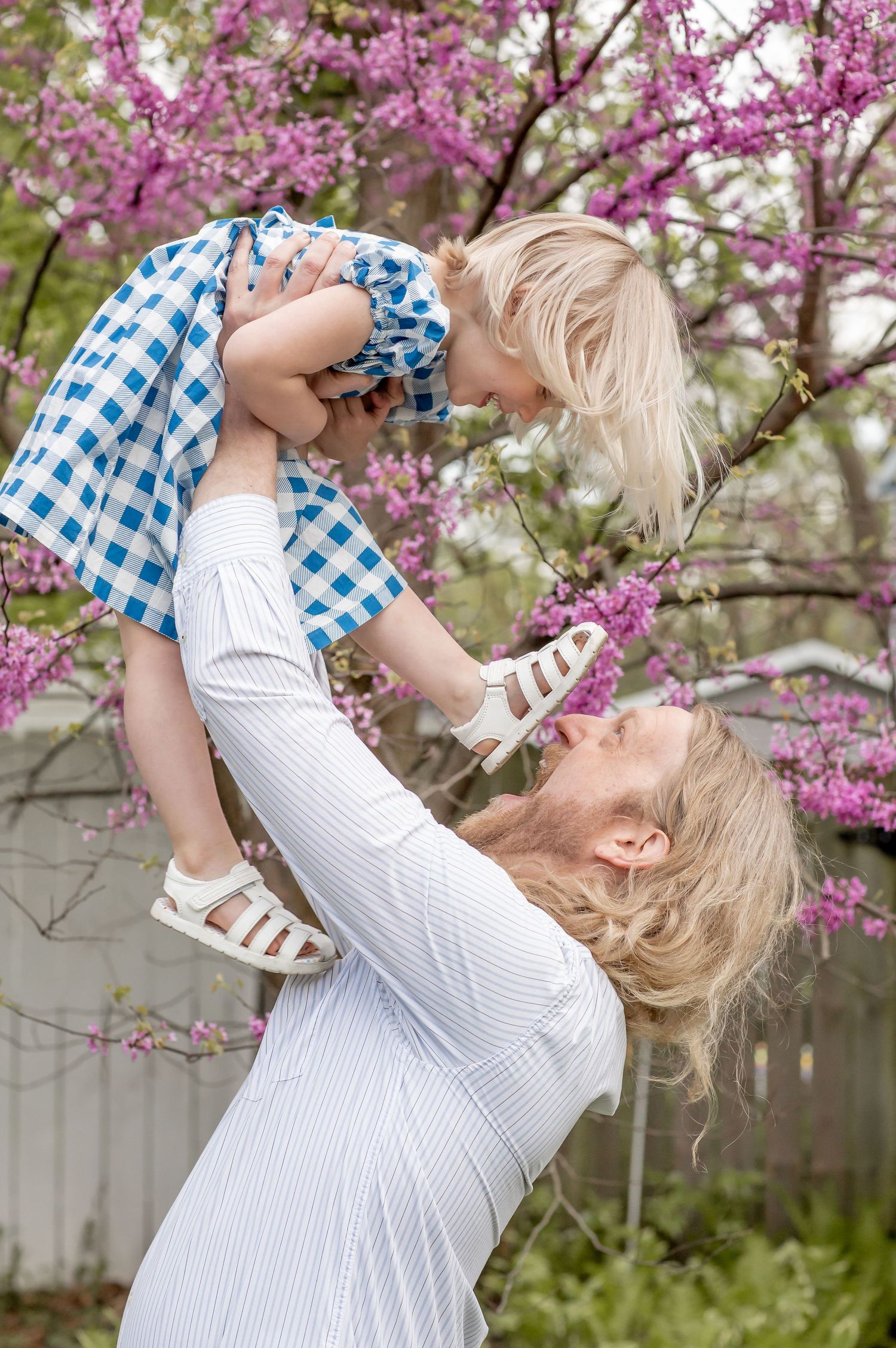 Mother lifting laughing toddler in blue gingham dress among blooming pink redbud trees in spring.