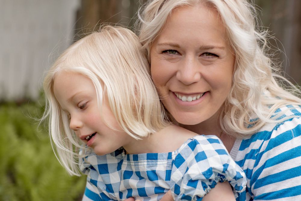 Smiling blonde mother hugging young daughter in matching blue striped outfits outdoors.