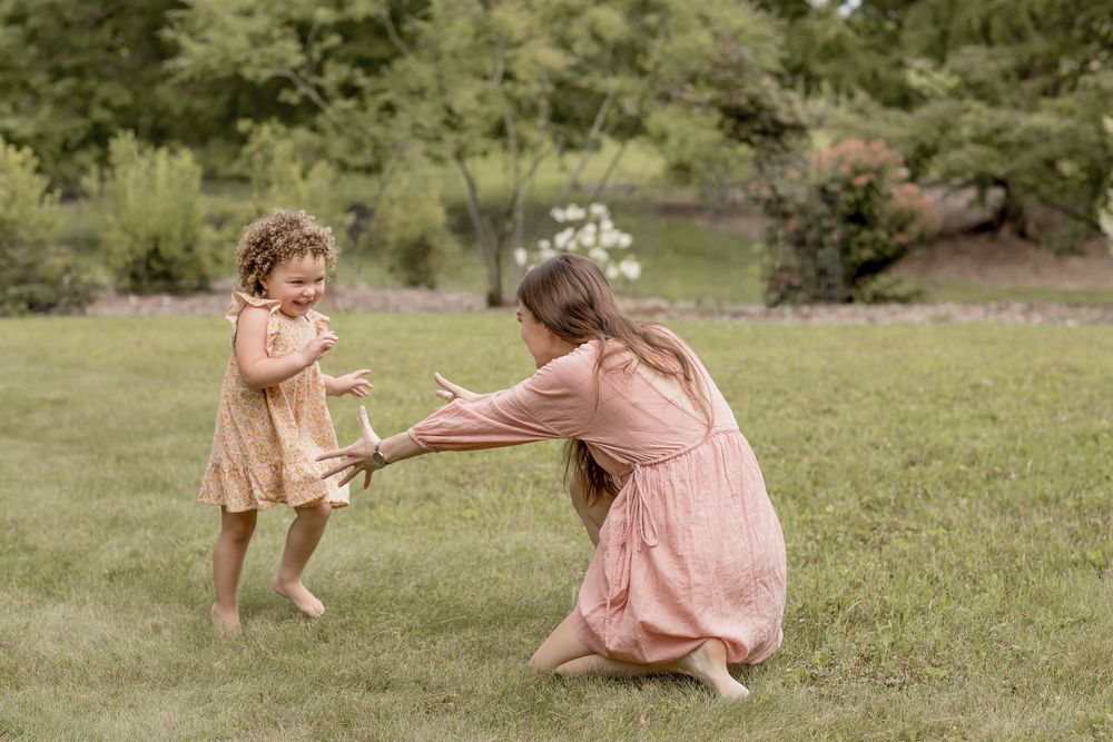 A mother in a pink dress kneels on grass, reaching out to her laughing toddler daughter outdoors.