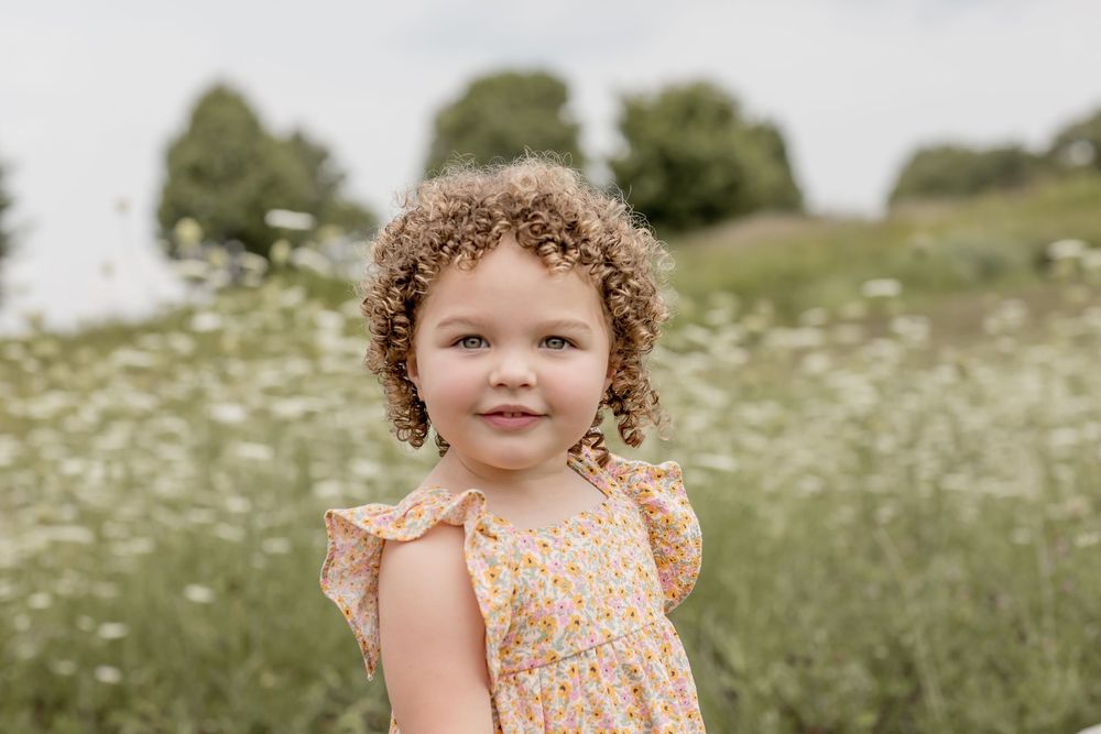 A curly-haired toddler girl in a yellow floral dress smiles in a wildflower meadow.
