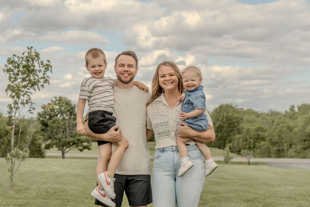 Happy family of four posing outdoors on green grass with cloudy sky, parents holding two young children.