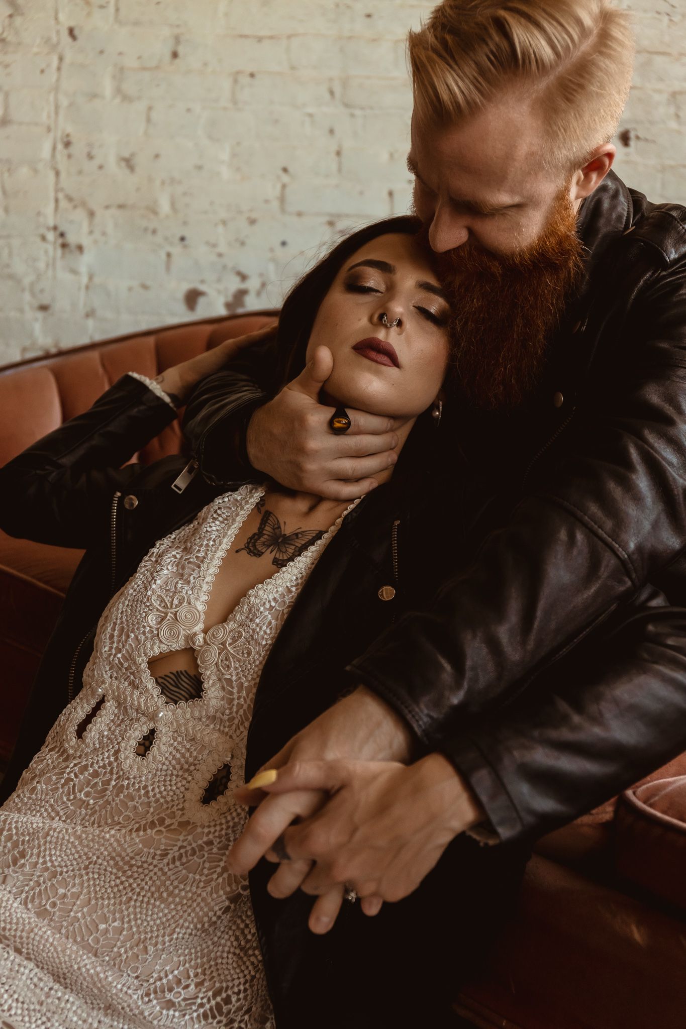A dramatic romantic scene with a couple in vintage attire lounging on a leather couch against a rustic brick wall.