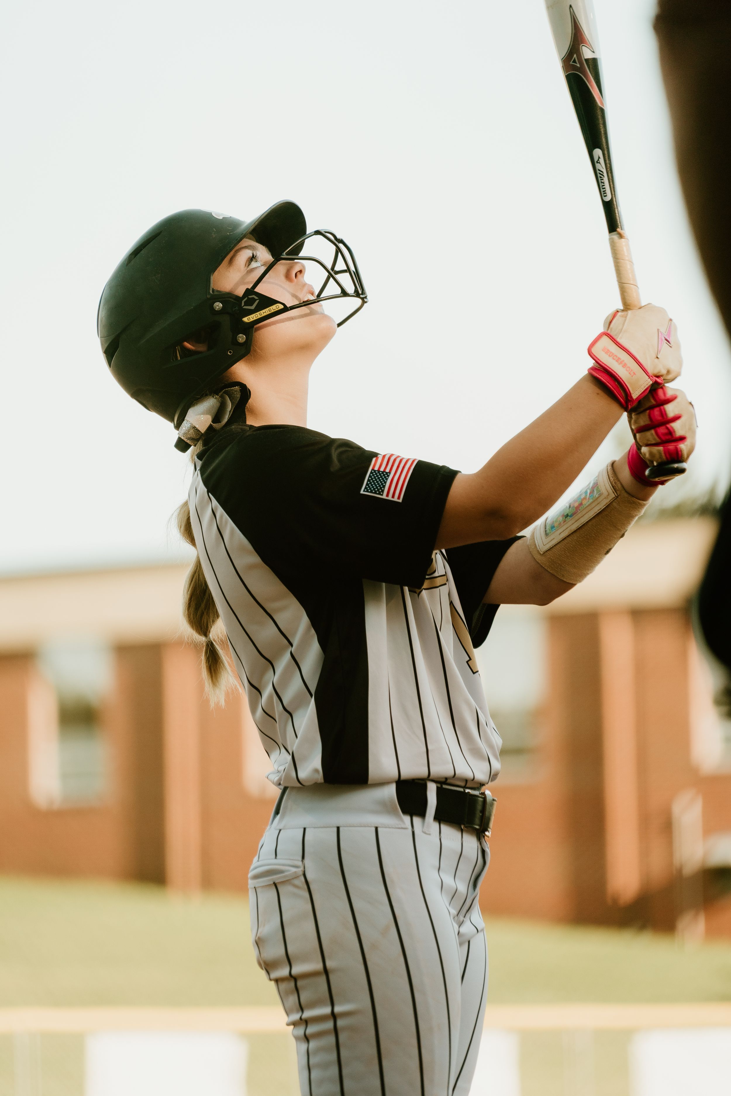 Female softball player in black helmet and pinstripe uniform swings bat during a game, wearing an American flag patch.