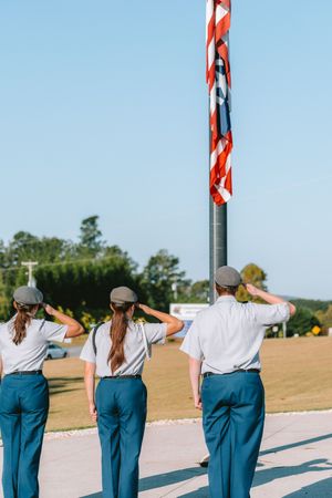 Three uniformed students salute flag during outdoor ceremony.