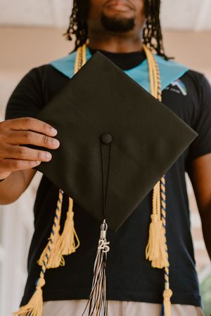 Graduation cap with gold tassel being held up against light background.
