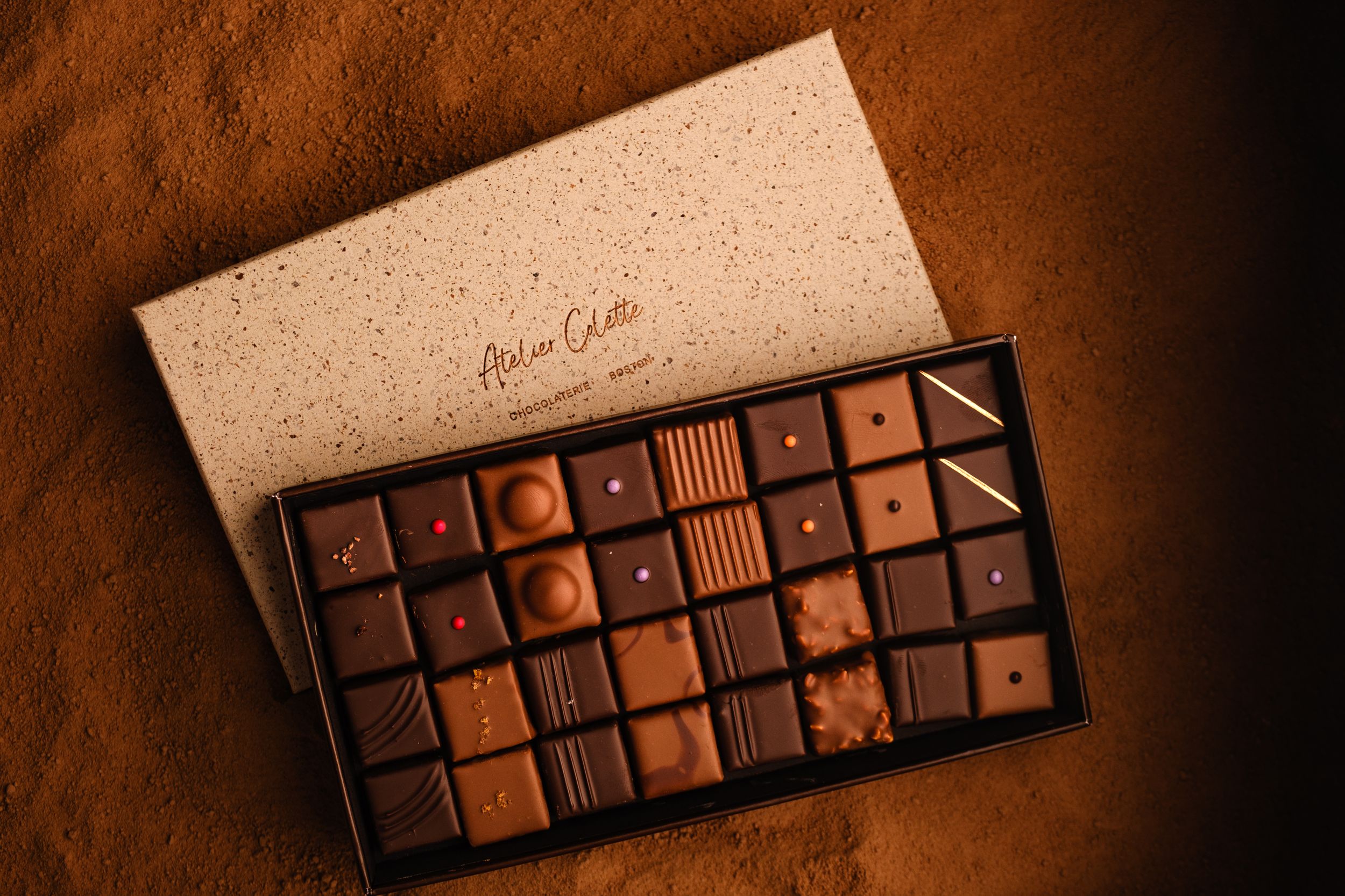 Luxury assorted chocolates in an elegant gift box by Anton Carte, displayed on rich cocoa powder background.