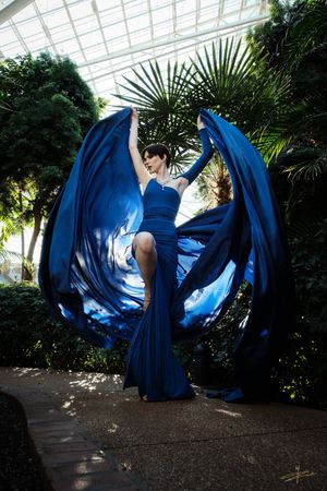 Woman in flowing blue gown poses dramatically indoors, fabric billowing like wings against lush tropical greenery.