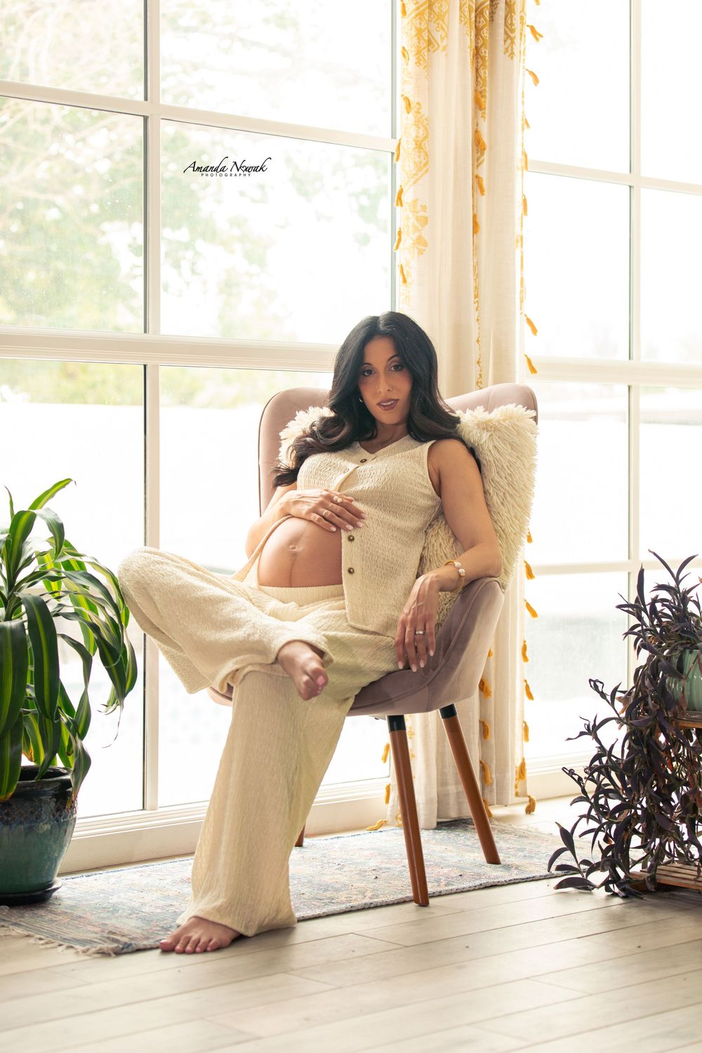 Pregnant woman in beige outfit sitting gracefully in a chair by a bright window surrounded by green plants.