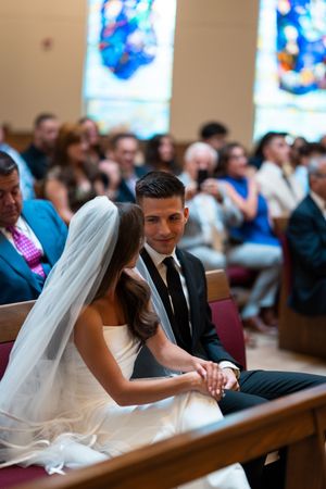 Couple sits in church pews during wedding ceremony with stained glass windows visible.