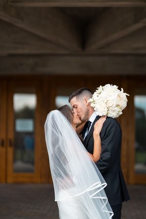 Romantic wedding couple sharing an intimate moment under a covered walkway with flowing veil and white bouquet.