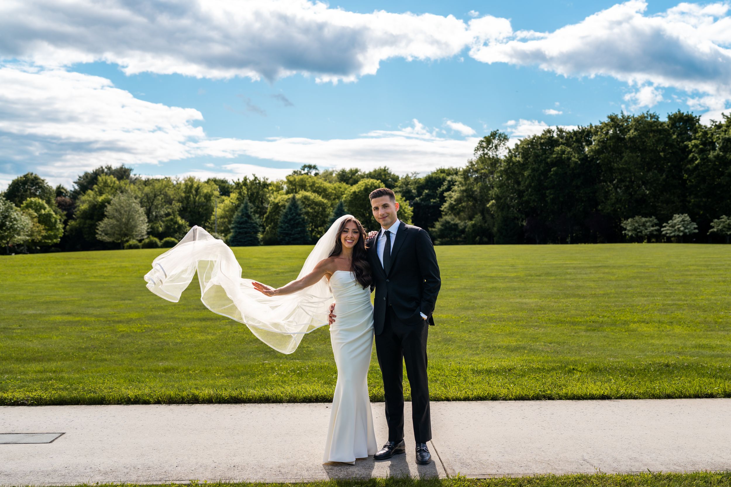 A couple poses in an outdoor field with a flowing white veil creating romantic movement in the breeze.