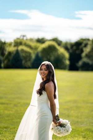 Bride in white dress with flowing veil poses joyfully in sunny green field.