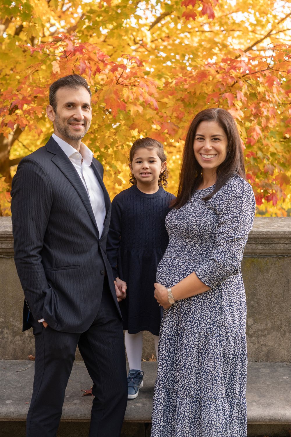 Family portrait taken outdoors with vibrant autumn foliage in the background.