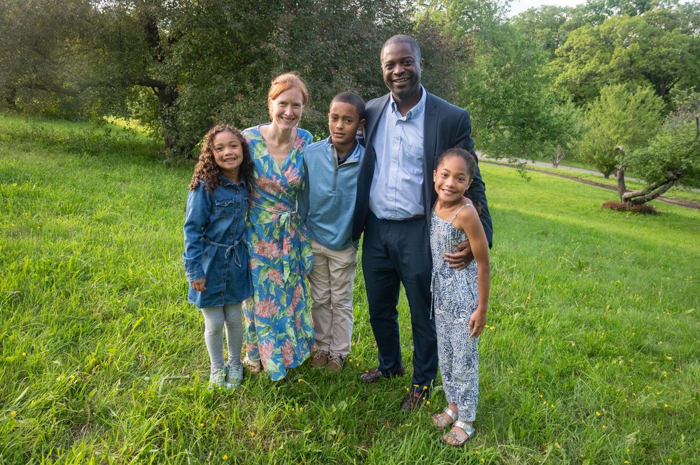 A joyful family of five poses together on a lush green lawn surrounded by trees on a warm evening.