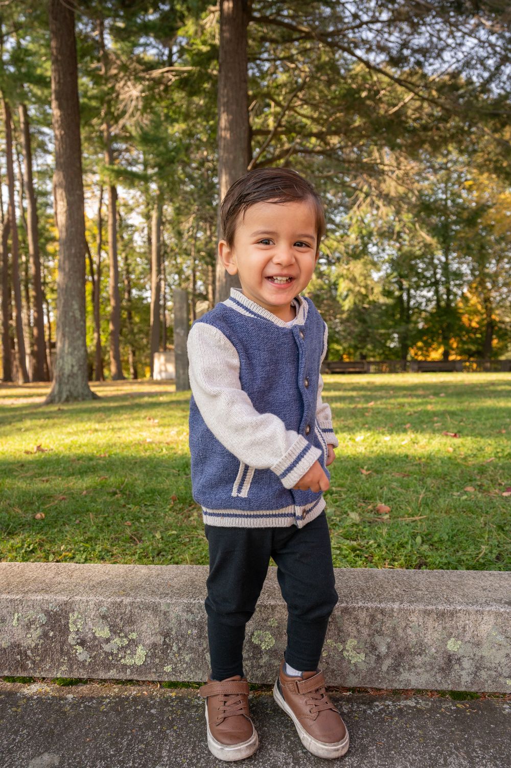 Small child wearing a blue sweater vest and jeans poses outdoors in a sunny park with autumn trees in background.
