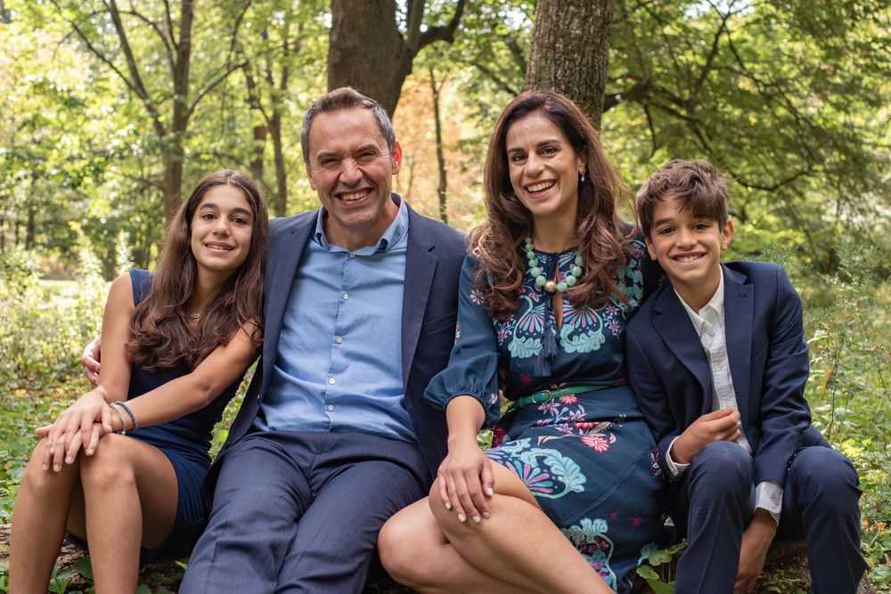 A group sits together outdoors on a wooden bench surrounded by lush green trees in a park setting.