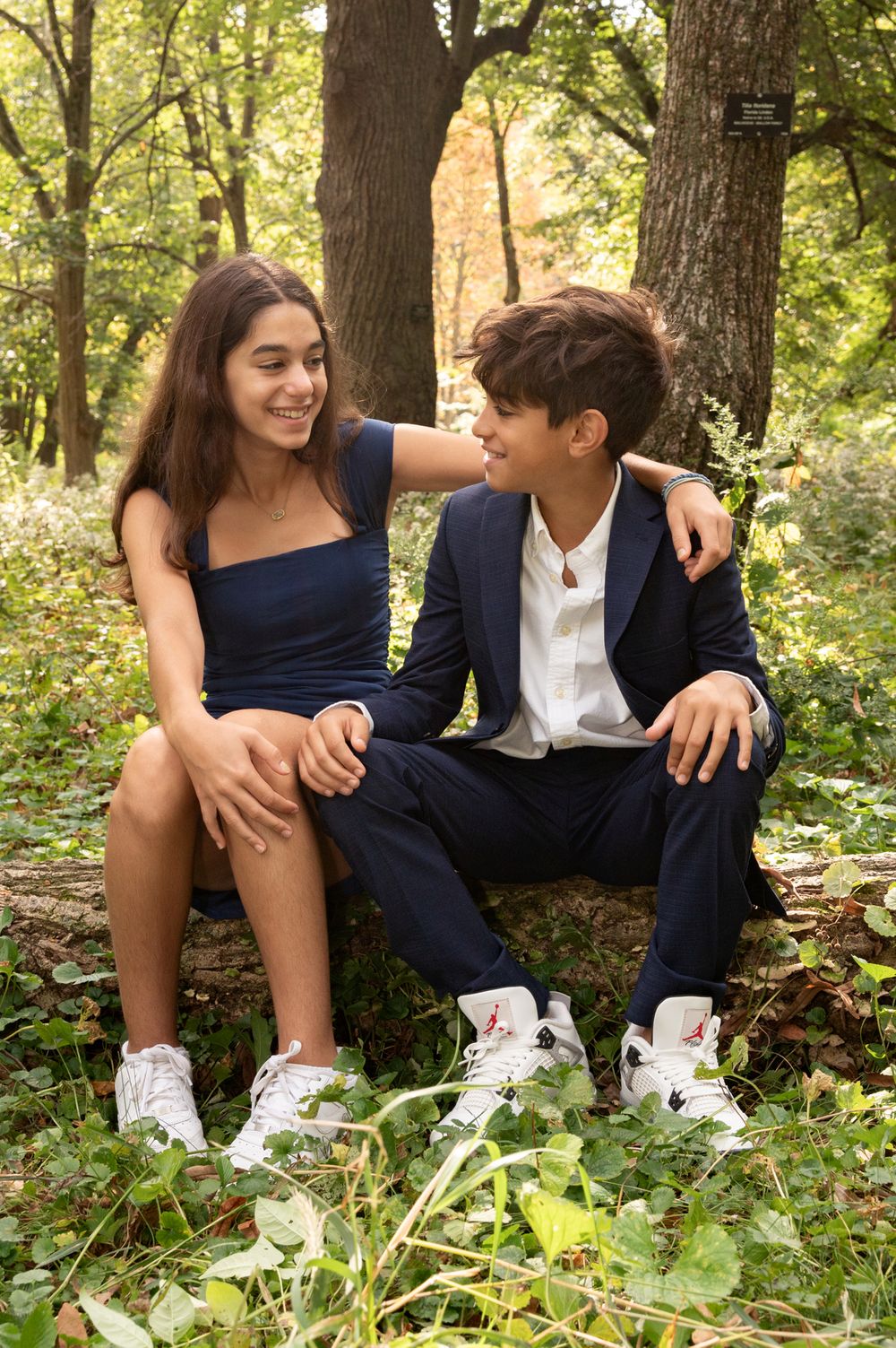 A couple in casual attire sits together on fallen logs in a lush forest setting.