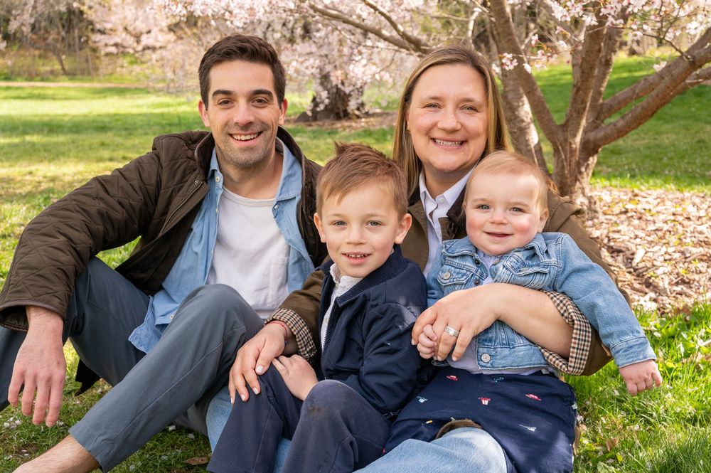 Family of four smiling outdoors under blooming cherry blossom tree on green grass in spring.