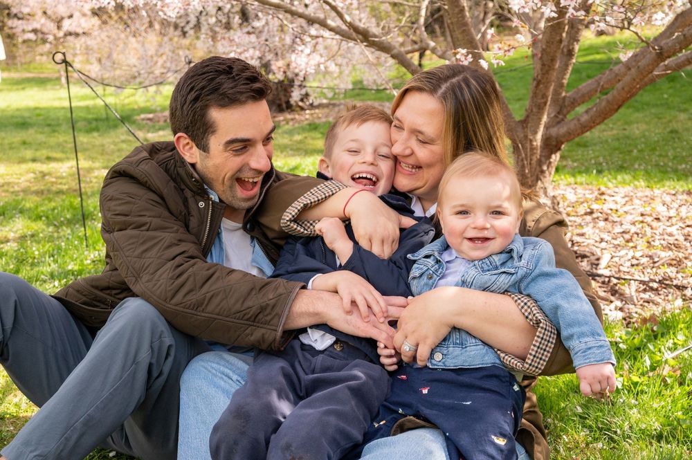 Happy family of four laughing together outdoors under blooming cherry blossom tree on green grass.