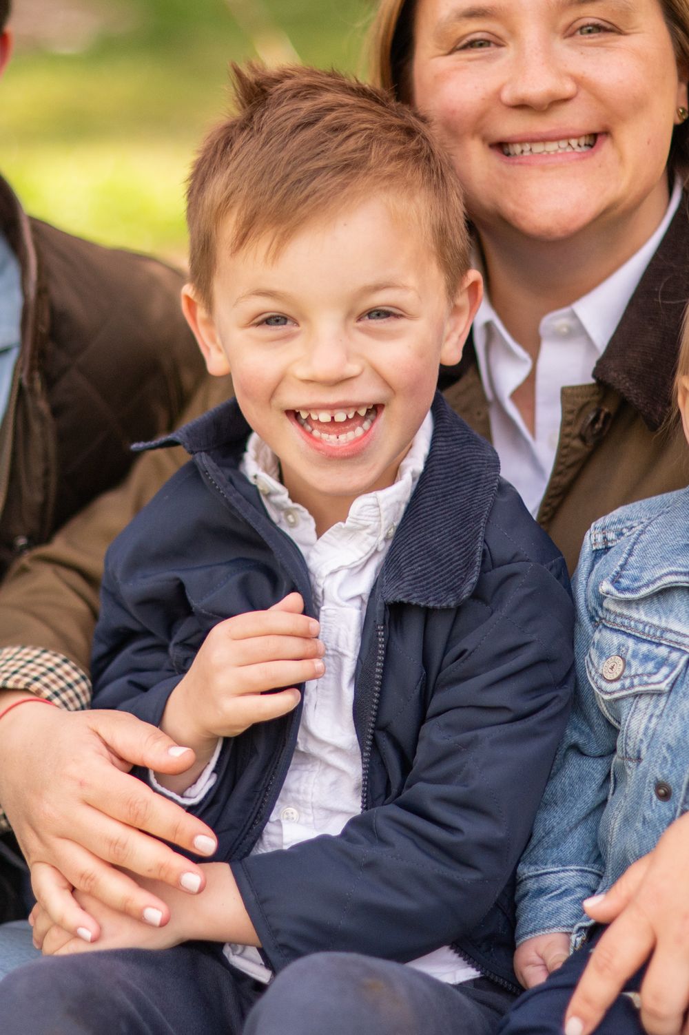 A joyful young boy with missing teeth smiles broadly, sitting with his family outdoors in a park setting.