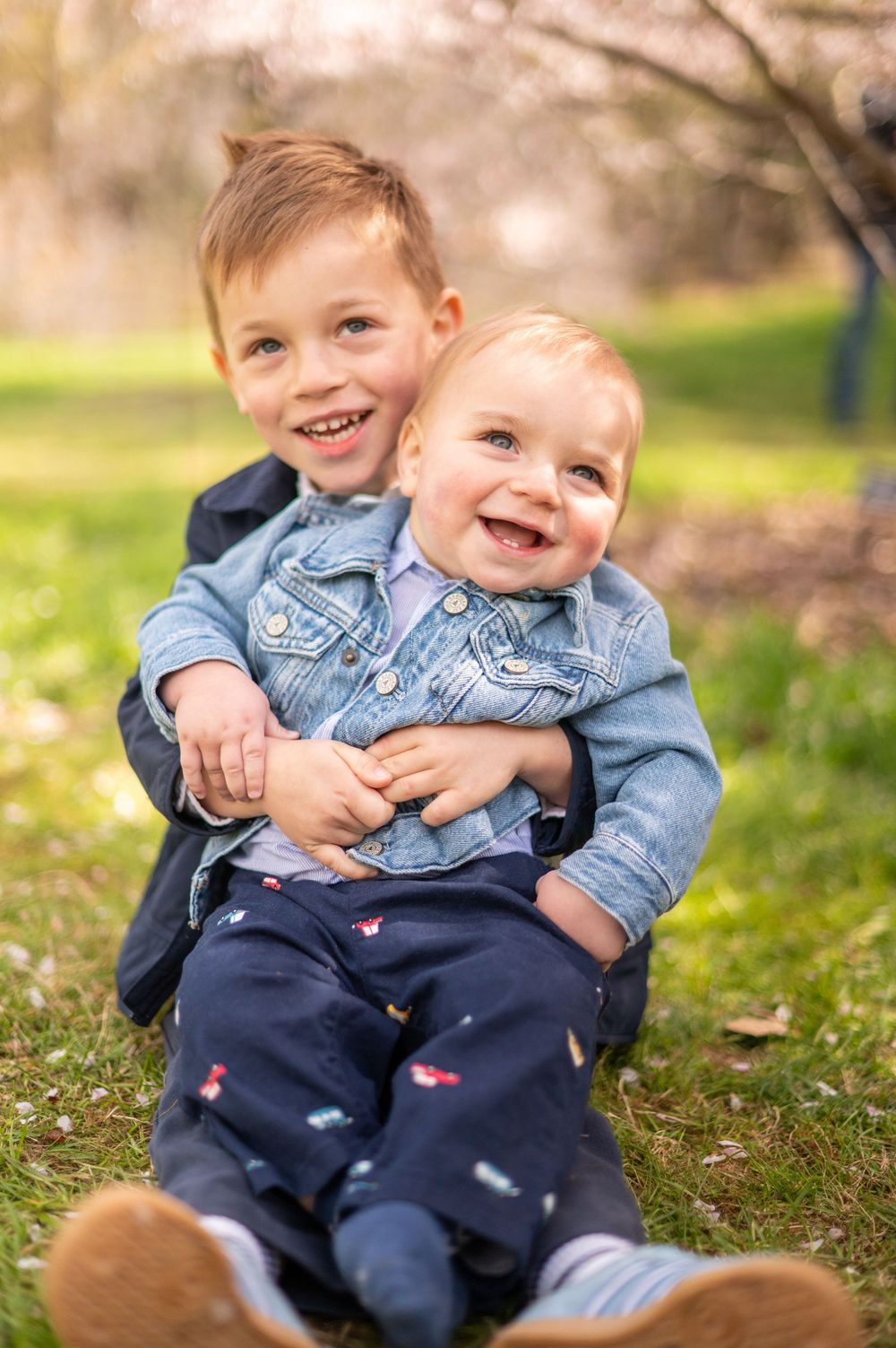 Two smiling young brothers in denim jackets sit together on green grass with spring blossoms in the background.