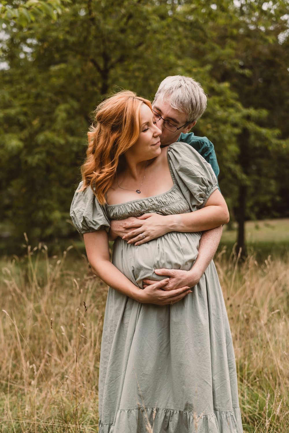 A couple embraces tenderly in a sunlit meadow with tall grass and trees in the background.