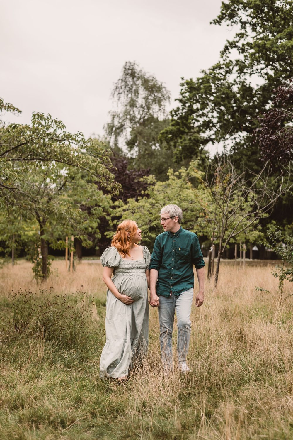 A couple walks together through a serene meadow with tall grass and trees on a sunny summer day.