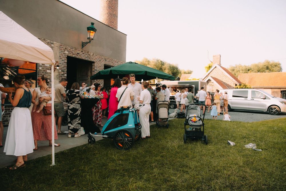 Outdoor gathering with guests socializing on a lawn near a rustic building, with strollers and a green umbrella visible.