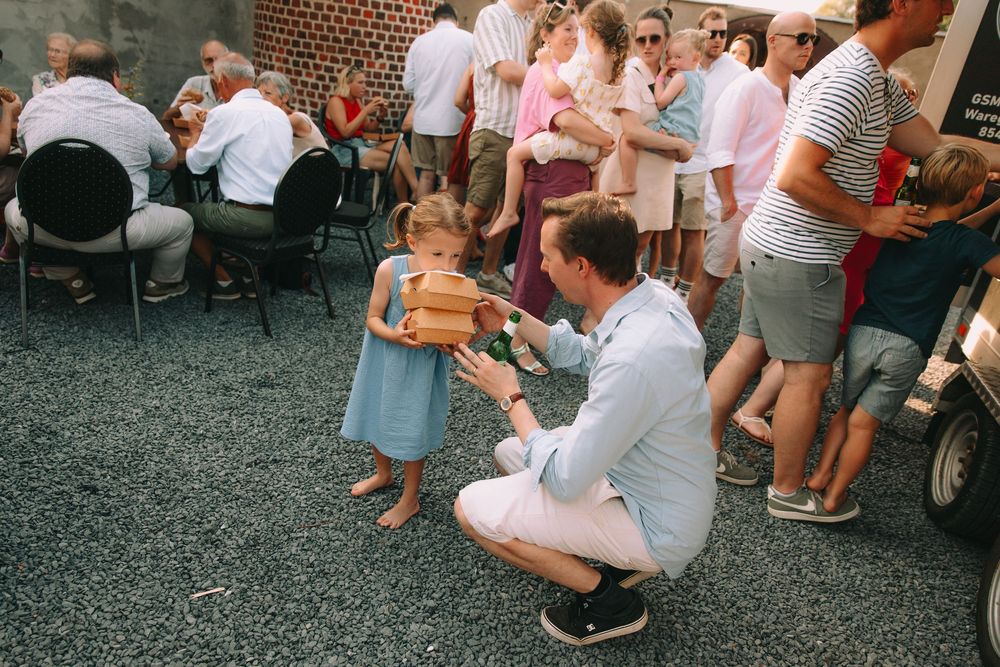 Man crouching to give a wooden gift to a young girl in a blue dress at an outdoor summer gathering.