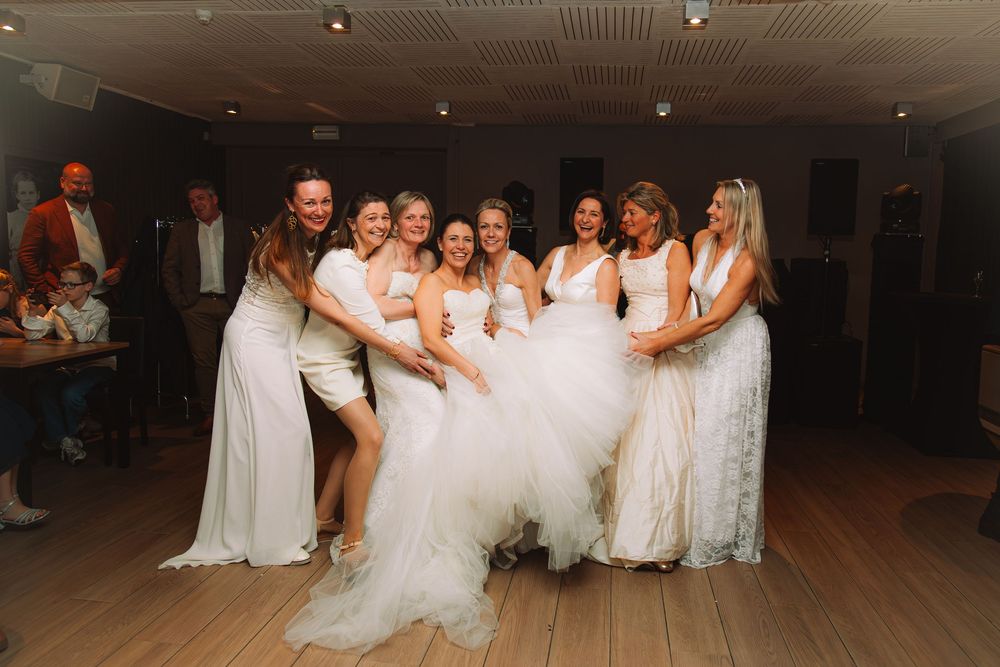 Bride surrounded by bridesmaids in white dresses, laughing and posing together on a wooden dance floor at a wedding reception.