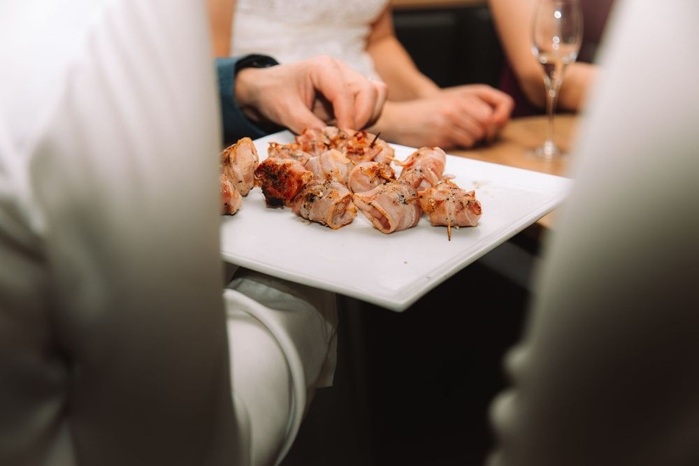 People sharing a plate of grilled meat skewers at a restaurant table with wine glasses in the background.