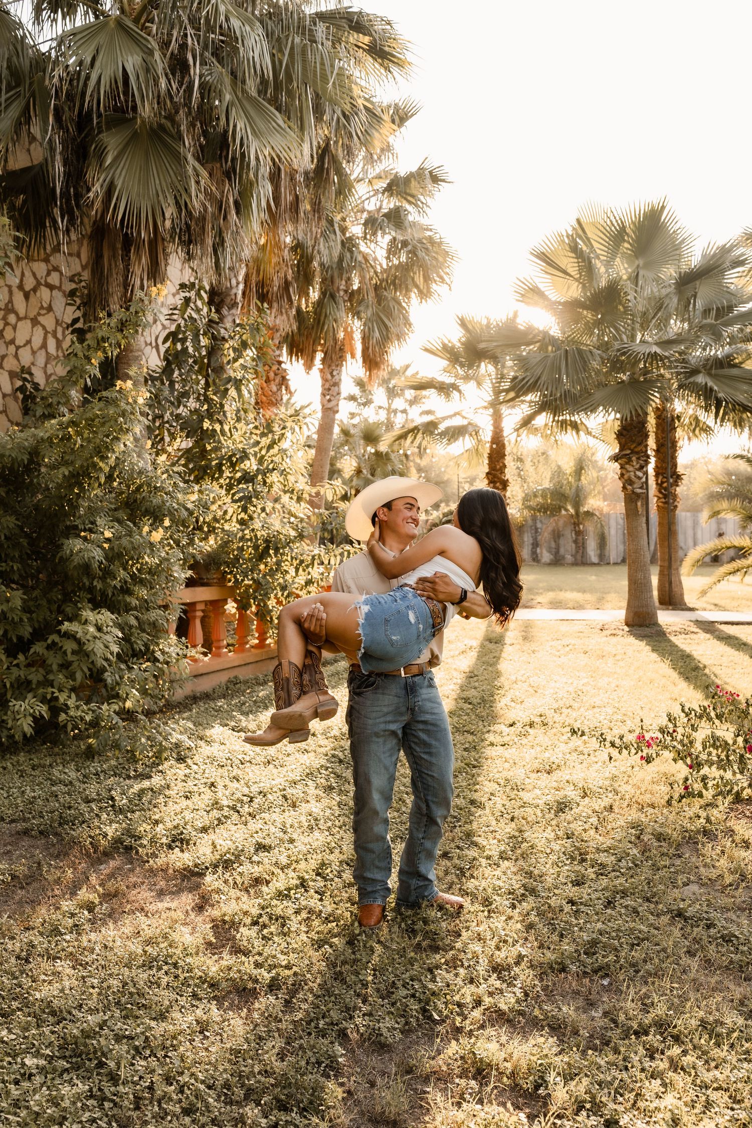 Couple sharing a romantic kiss while dancing on lawn in front of a stone villa with palm trees.