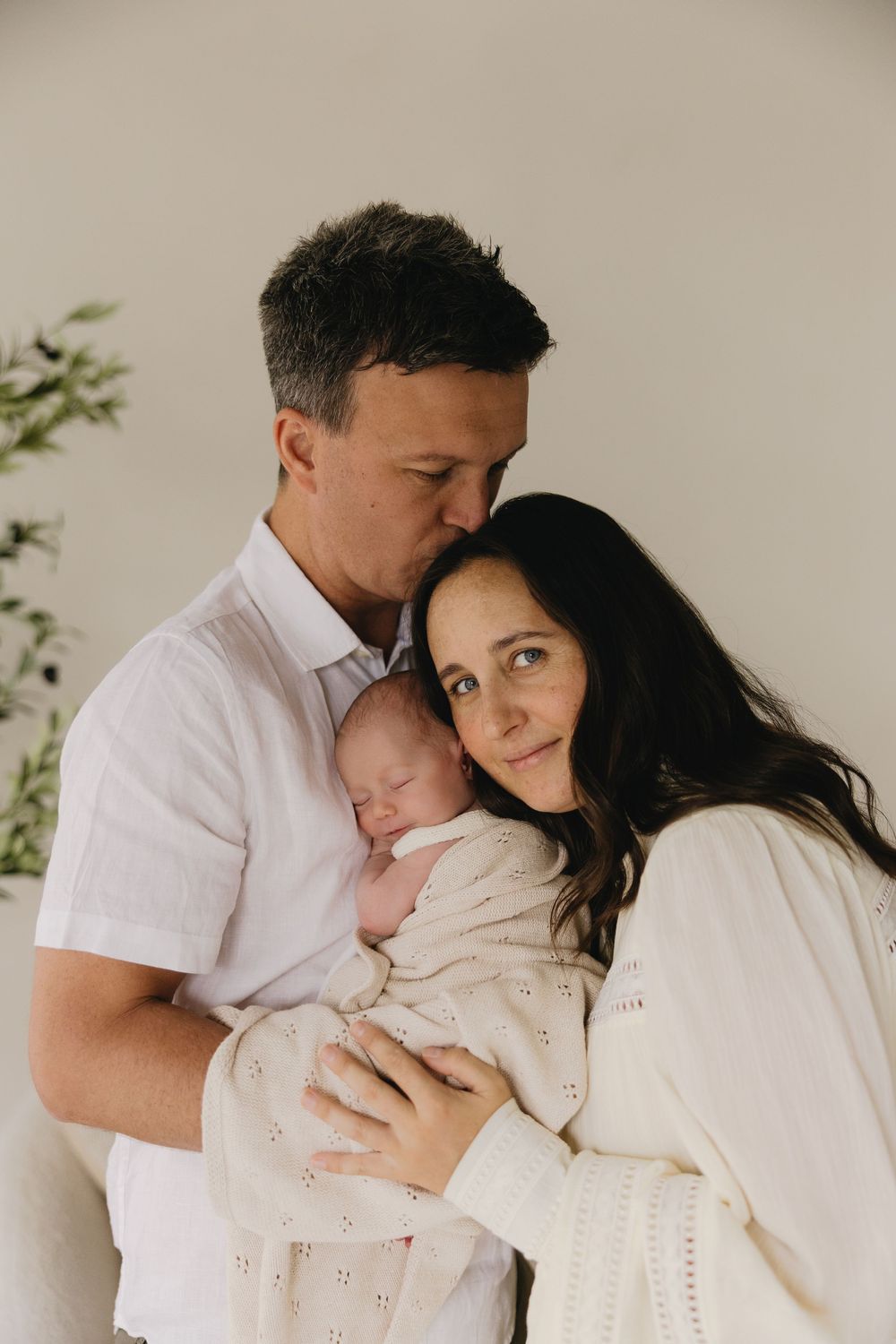 A tender family moment captured in soft tones as parents embrace their newborn baby in matching white outfits.